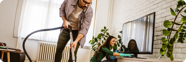 A young Caucasian man hoovering in a living room and a happy looking young Caucasian woman cleaning a TV stand in the background.  